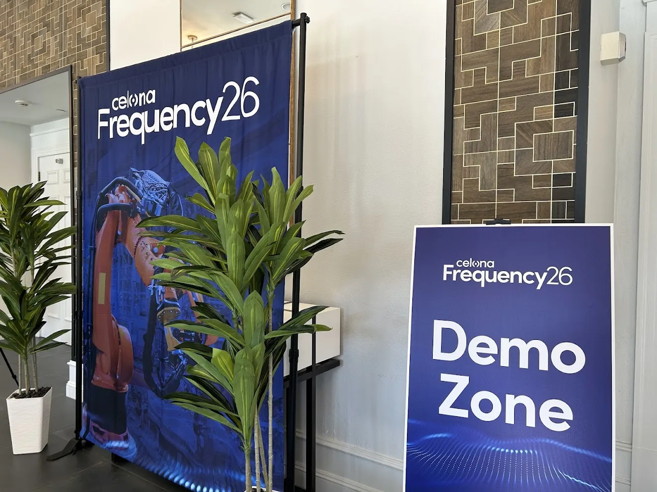 Tall blue Celona Frequency26 banner beside a purple Demo Zone sign, with leafy potted plants in front.
